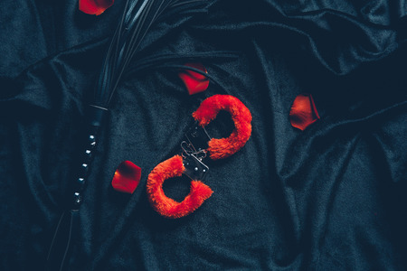 top view of red fluffy handcuffs, leather whip and rose petals on black fabricの写真素材