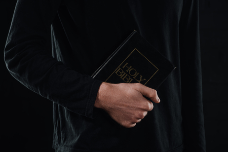 cropped shot of priest holding holy bible isolated on blackの写真素材
