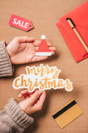 cropped image of woman holding merry christmas lettering and santa hat over surface with credit card and saleの写真素材