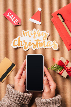 cropped image of woman holding smartphone with blank screen over surface with credit card, gift box and merry christmas letteringの写真素材