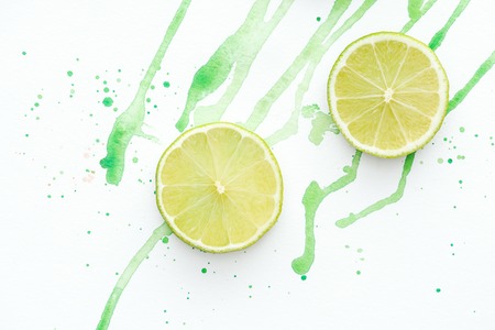 elevated view of two pieces of ripe limes on white surface with green watercolorの写真素材