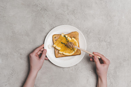 top view of human hands with honey dipper and healthy sandwich with orange slices on greyの写真素材