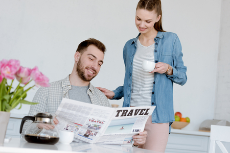 husband reading travel newspaper while smiling wife with coffee standing near in kitchenの写真素材