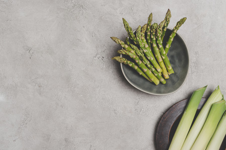 top view of fresh organic asparagus and leeks on plates on greyの写真素材