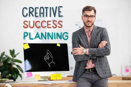 portrait of businessman in suit and eyeglasses standing at workplace with computer in office, creative success planning inscriptionの写真素材
