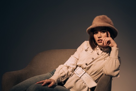 portrait of beautiful young woman in fashionable clothing and hat looking at camera while sitting in armchairの写真素材