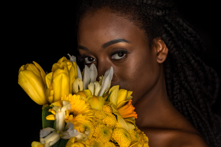beautiful young african american woman holding yellow flowers and looking at camera isolated on blackの写真素材