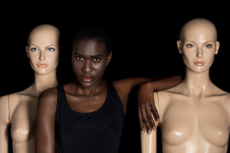 attractive young african american woman in bodysuit standing between mannequins and looking at camera on blackの写真素材