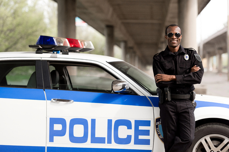 handsome african american police officer with crossed arms leaning back on car and looking at cameraの写真素材