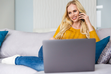 beautiful smiling blonde girl talking by smartphone and using laptop at homeの写真素材
