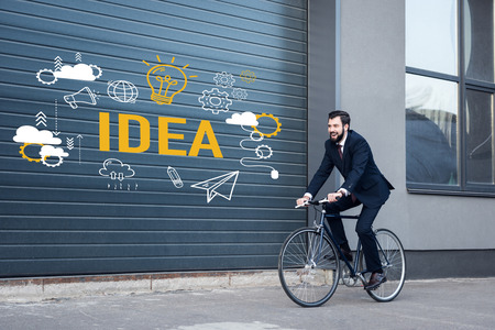 smiling young businessman in suit riding bicycle on street with idea inscription and business icons on gate entranceの写真素材