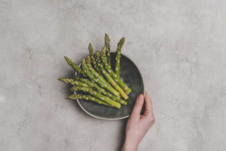 partial top view of person holding plate with fresh healthy asparagus on greyの写真素材