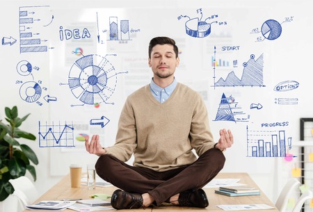 relaxed businessman with eyes closed sitting in lotus position on table in office with business graphs aroundの写真素材