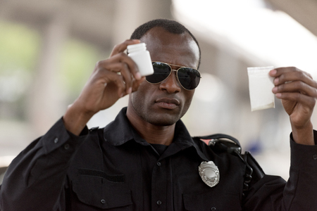 african american policeman holding drugs in plastic zipper and pills jarの写真素材