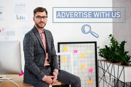portrait of young businessman in suit and eyeglasses sitting on table in office, advertise with us inscriptionの写真素材