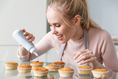 beautiful smiling young woman in apron preparing delicious muffins with jamの写真素材