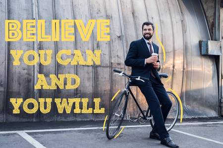 smiling businessman with disposable cup of coffee leaning on bicycle on street with believe you can and you willの写真素材