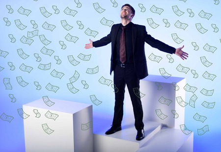 businessman in black suit with outstretched arms standing on white block with falling dollar...