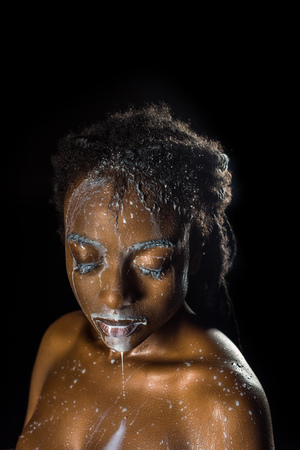 seductive african american girl with milk drops on face and body posing with closed eyes isolated on blackの写真素材