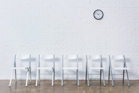 Row of empty chairs by white brick wall with clockの写真素材