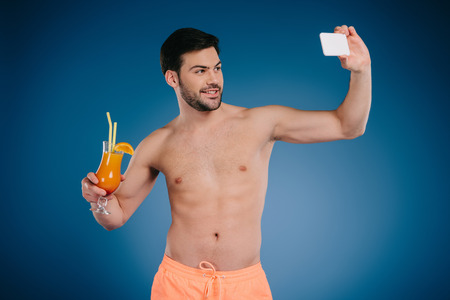 Handsome shirtless young man in shorts holding glass of summer cocktail and taking selfie with smartphone on blue backgroundの写真素材