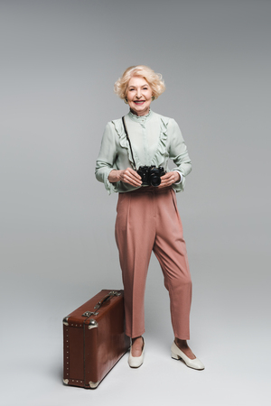 beautiful senior woman with vintage suitcase and film camera on greyの写真素材