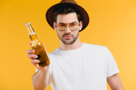 Handsome young man in hat and sunglasses holding bottle with summer drink and looking at camera isolated on yellow backgroundの写真素材