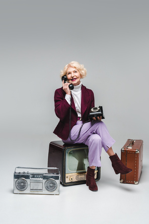 beautiful senior woman talking by phone while sitting on vintage tv on greyの写真素材