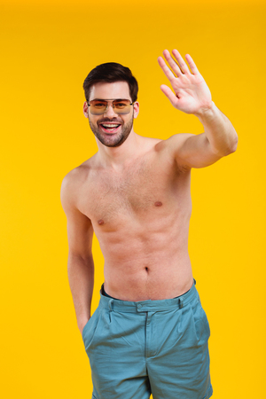 Handsome shirtless young man in shorts and sunglasses waving hand and smiling at camera isolated on yellow backgroundの写真素材