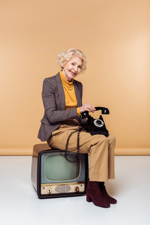 Smiling stylish senior woman with rotary phone sitting on vintage televisionの写真素材