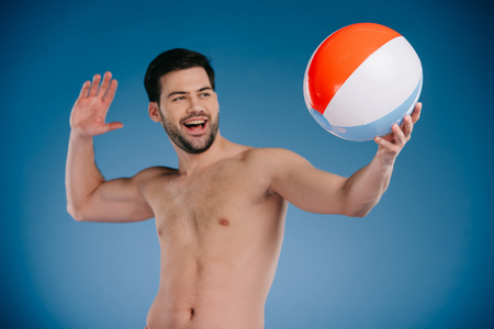 Smiling young bare chested throwing beach ball and looking away on blue backgroundの写真素材
