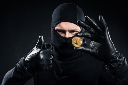 Man in black balaclava holding bitcoin and showing thumb upの写真素材