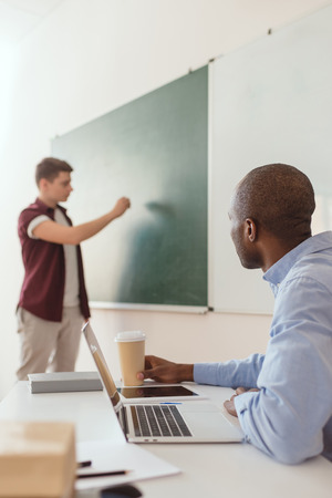 High school student writing at chalk board and African American teacher sitting at desk with laptop and coffee cupの写真素材