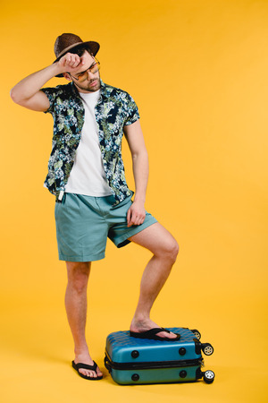 Young exhausted male tourist standing on suitcase isolated on yellow backgroundの写真素材