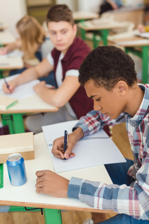 Elevated view of African American teenage schoolboy writing in textbook and classmates on behindの写真素材