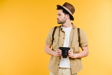Young man in hat with backpack holding camera and looking away isolated on yellow backgroundの写真素材