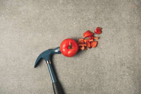 Flat lay with cut fresh tomato, tomato pieces and hammer on grey concrete tabletopの写真素材