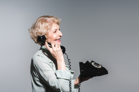 Side view of smiling senior woman talking by vintage phone isolated on grey backgroundの写真素材