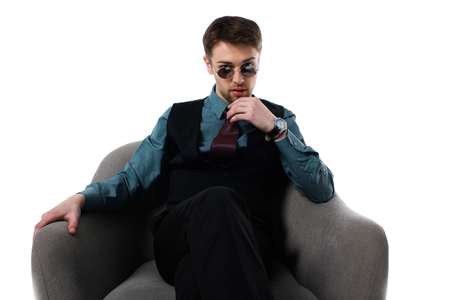 Portrait of pensive stylish spy in sunglasses sitting in armchair isolated on white backgroundの写真素材