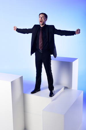 Businessman in black suit with outstretched arms standing on white block isolated on blue backgroundの写真素材