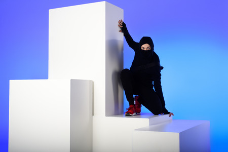 Ninja in black clothing with katana behind standing on white block isolated on blue backgroundの写真素材