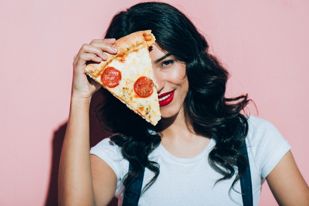 Obscured view of beautiful smiling woman covering eye with piece of pizza on pink backgroundの写真素材