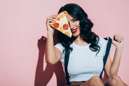 Obscured view of beautiful smiling woman covering eye with piece of pizza on pink backgroundの写真素材