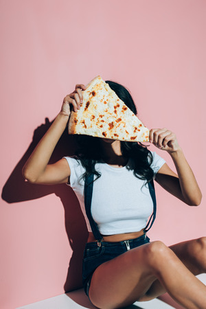 Obscured view of woman covering face with piece of pizza on pink backgroundの写真素材