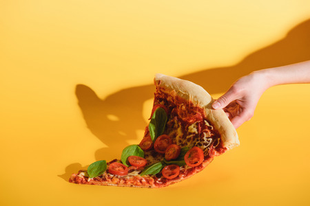 Partial view of woman holding piece of pizza with cherry tomatoes in hand on yellow backgroundの写真素材