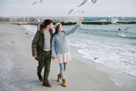 Young couple walking on winter sea shore, girl pointing at seagullsの写真素材