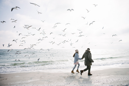 Young couple holding hands and walking on winter sea shore and looking at seagulls