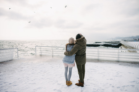 Back view of young couple embracing on winter quay and looking at the seaの写真素材
