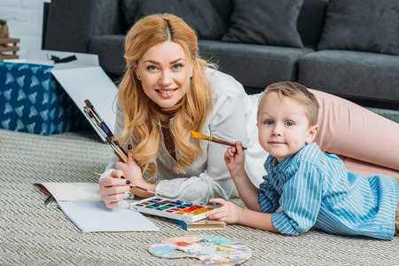 Little boy and mother laying on rug and holding paintbrushes in hand