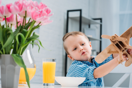 Little boy playing with wooden airplane at table with plate, juice and tulipsの写真素材